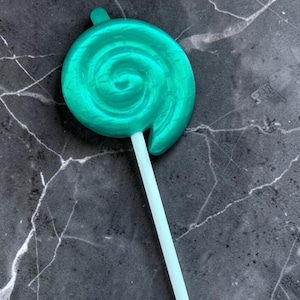 May include: A teal-colored, swirled lollipop with a white stick. The candy has a glossy finish and a spiral design. The lollipop is set against a dark gray, marble-patterned background.