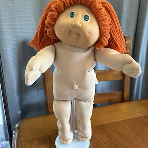 Puede incluir: Una muñeca vintage con un cuerpo suave color carne y una cara redonda. La muñeca tiene ojos verdes brillantes, una nariz pequeña y una ligera sonrisa. Su pelo de hilo naranja está peinado en capas. La muñeca está sobre una base blanca.