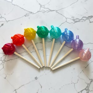 May include: A collection of seven colorful lollipops arranged in a semi-circle. Each lollipop features a different color, including red, orange, yellow, green, blue, purple, and pink. The lollipops are on wooden sticks, set against a marble background.