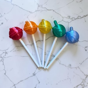 May include: Five round lollipops arranged in a fan shape, each featuring a different translucent color including red, orange, yellow, green, and blue. Each lollipop has a smooth, spherical head attached to a thin, straight white plastic stick.