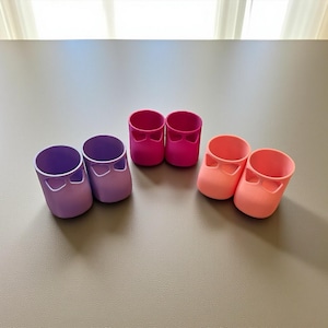 Puede incluir: Seis pequeños recipientes de plástico con forma de zapato en tonos morado, rosa y melocotón. Cada uno tiene un recorte en forma de lazo. Los recipientes están dispuestos sobre una superficie neutra.