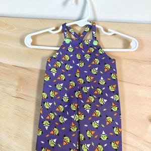 Può includere: Una salopette viola con un motivo ripetuto di streghe dal volto verde con cappelli arancioni e bianchi, intervallato da piccole caramelle bianche. Il capo presenta due spalline fissate sul davanti con bottoni di colore verde brillante.