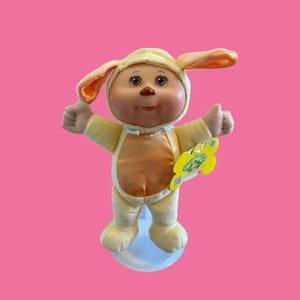 Puede incluir: Muñeca Cabbage Patch Kids con un disfraz de conejito amarillo con vientre naranja claro. La muñeca tiene ojos marrones y hace un gesto de pulgar hacia arriba con ambas manos. Una etiqueta amarilla está adherida al atuendo.