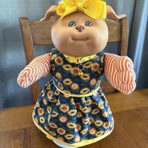 Puede incluir: Una muñeca de perro de peluche con un lazo amarillo, que lleva un vestido azul marino con un estampado de girasoles. La muñeca tiene mangas a rayas naranjas y blancas y un collar amarillo. La muñeca está sobre una superficie de madera.