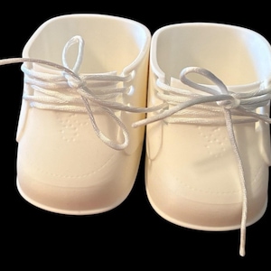 May include: A pair of cream-coloured baby shoes with white laces, presented on a black backdrop. The shoes feature a classic trainer design, a rounded toe, and a textured pattern on the front.