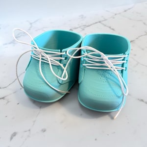 May include: A pair of turquoise baby shoes with white laces. The shoes are made of a solid material and have a classic sneaker design. The laces are tied in a bow, and the shoes are sitting on a white marble surface.