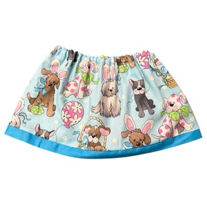 Puede incluir: Falda azul claro con un estampado repetido de perros de dibujos animados con orejas de conejo, huevos de Pascua y globos. La falda tiene un ribete azul y una cintura fruncida. La falda mide unos 30 cm de largo.