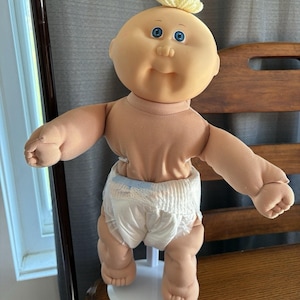 Puede incluir: Una muñeca bebé vintage con una cara redonda, ojos azules y un mechón de pelo amarillo. La muñeca lleva un pañal y está de pie sobre un soporte blanco. Los brazos de la muñeca están extendidos.
