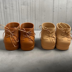May include: Two pairs of miniature ankle boots with rounded toes and thin laces. One pair is a deep tan colour, and the other pair is a lighter beige colour. Each boot features visible stitching along the eyelet panels and a smooth, matte finish.