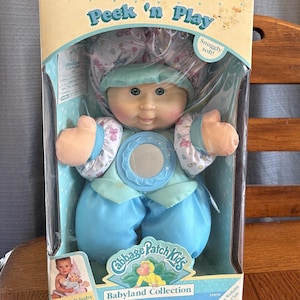 May include: A Cabbage Patch Kids Babyland Collection doll in its original packaging. The doll is wearing a blue outfit and a matching bonnet. The box reads "Peek 'n Play" and "So-soft baby with a play surprise!"