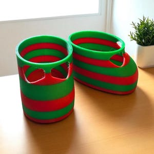 May include: Two 3D-printed decorative containers, one cylindrical and one boot-shaped, both with alternating horizontal stripes of red and green. The cylindrical container has two cut-out shapes. The boot-shaped container has a rounded toe.