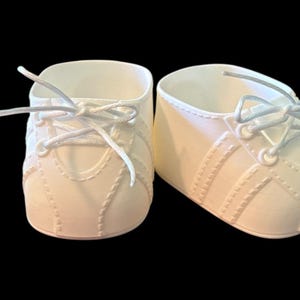 Może przedstawiać: Para kremowych bucików dla niemowląt ze sznurowadłami, na czarnym tle. Buty mają klasyczny design sneakersów z przeszytymi detalami i teksturowanym wyglądem. Sznurowadła są zawiązane.