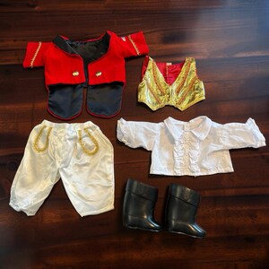 May include: A doll clothing set including a red jacket with gold trim and two buttons, a gold metallic vest with red accents, white satin pants with gold pocket trim, a white ruffled long-sleeve shirt, and a pair of black knee-high boots.