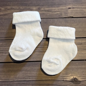 May include: Two pairs of white baby socks with folded-over cuffs. The socks are made of a soft, ribbed knit material. The socks are displayed on a wooden surface.