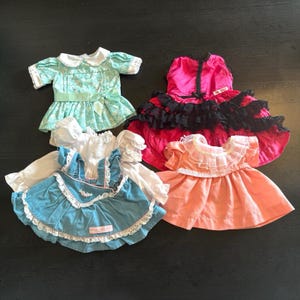 Puede incluir: Cuatro vestidos de muñeca de varios estilos y colores se muestran sobre una superficie oscura. Los vestidos incluyen un vestido floral azul claro con un lazo, un vestido rosa con encaje negro, un vestido azul con encaje blanco y un vestido melocotón con cuello con volantes.