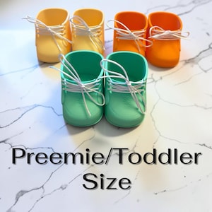 Op de afbeelding: Zes kleine schoenen in pastelgeel, feloranje en mintgroen, elk voorzien van witte veters. De schoenen zijn in paren gerangschikt en de tekst Preemie/Toddler Size staat onderaan de afbeelding. De schoenen hebben een gladde afwerking.