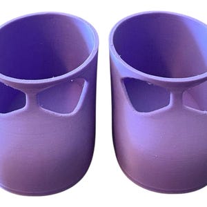 Puede incluir: Dos macetas en forma de zapato, impresas en 3D, de color lavanda. Cada maceta tiene una base redondeada y una sección superior cilíndrica con un diseño recortado que se asemeja a la pala de un zapato. Las macetas están diseñadas para contener pequeñas plantas o flores.