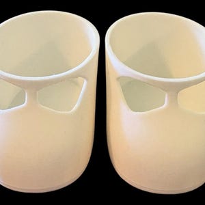 Może przedstawiać: Dwa małe, kremowe buty z wyciętym wzorem z przodu. Buty mają gładką powierzchnię i minimalistyczny styl. Tło jest czarne. Buty są prawdopodobnie dla dziecka.