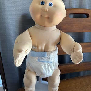 Puede incluir: Muñeca Cabbage Patch Kid vintage con cuerpo de tela suave y cabeza de vinilo. La muñeca tiene ojos azules, expresión sonriente y lleva un pañal. Está sobre un soporte blanco.
