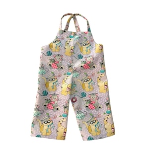 May include: Light purple baby overalls with a repeating pattern of cartoon cats wearing hats and holding yarn balls. The overalls have adjustable straps and button closures. The fabric is decorated with small white polka dots.