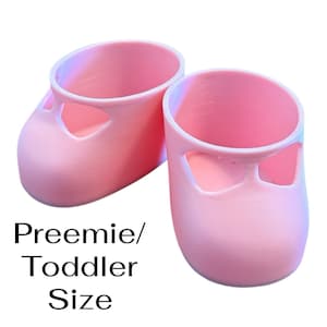 Op de afbeelding: Een paar lichtroze schoenen voor premature baby's/peuters. De schoenen hebben een ronde neus en een uitgesneden ontwerp aan de bovenkant. De tekst "Preemie/Toddler Size" staat eronder in het zwart.
