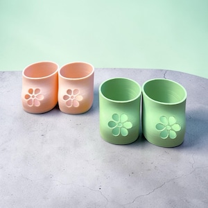 May include: Four ceramic flower-shaped candle holders in two colors. Two are peach-colored, and two are green. Each has a flower cut-out on the front. The candle holders are on a gray surface with a light green background.