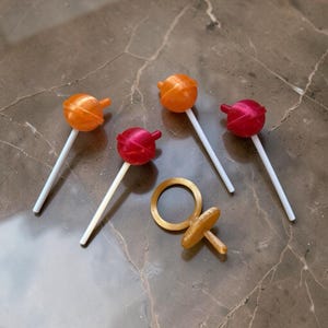 May include: Four lollipops with round, translucent heads in orange and red, each attached to a thin white stick. Beside them lies a gold-colored ring with a small, protruding circular attachment, resembling a pacifier or a decorative charm.