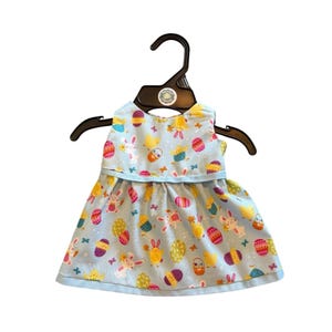 May include: A light blue baby dress with an Easter-themed print featuring bunnies, chicks, and decorated eggs. The dress is sleeveless and has a light blue trim. It is hanging on a black hanger.