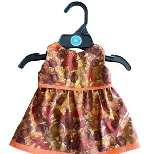 May include: A doll dress with a fall-themed leaf and acorn pattern, displayed on a black hanger. The dress features a brown, orange, and red color scheme with an orange trim. The dress is sleeveless and has a flared skirt.