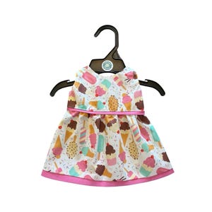 Puede incluir: Un vestido blanco sin mangas con un ribete rosa y una cinta rosa alrededor de la cintura. El vestido está decorado con coloridos conos de helado, polos y cupcakes. El vestido está colgado de una percha negra.