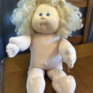 May include: A vintage Cabbage Patch Kid doll with a soft, pink body and a round face. The doll has blue eyes, a small mouth, and curly blonde hair. It is sitting on a wooden surface.