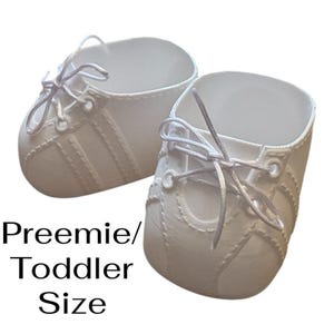 May include: A pair of white preemie/toddler shoes with white laces. The shoes have a classic sneaker design with stitched details and a rounded toe. The text "Preemie/Toddler Size" is displayed below the shoes.
