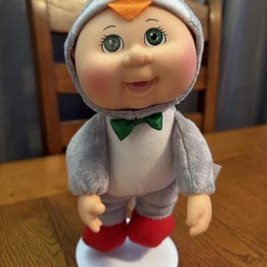 May include: A Cabbage Patch Kids doll dressed in a gray penguin costume with an orange beak and green bow tie. The doll has green eyes and red shoes, standing on a white base.