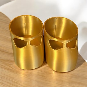 May include: Two gold-colored cylindrical containers with a unique design. Each container has a rounded top and a cut-out design on the front, giving them a distinctive appearance. The containers are made of a shiny, metallic material.