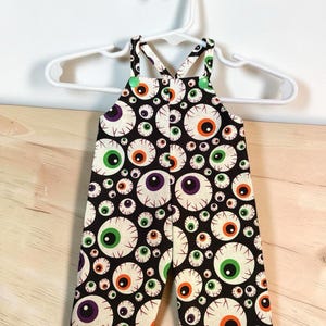 Op de afbeelding: Een tuinbroek met een zwarte achtergrond en een herhalend patroon van oogbollen in verschillende maten en kleuren, waaronder groene, oranje en paarse irissen. Het kledingstuk heeft twee schouderbanden die vastzitten met felgroene drukknopen.