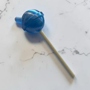 Könnte beinhalten: Ein blauer Lutscher mit einem runden Bonbonkopf und einem hellbraunen Stiel. Die Süßigkeit hat ein Wirbeldesign und ist vor einem weißen Marmorhintergrund platziert. Der Lutscher ist eine süße Leckerei.