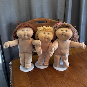 Könnte beinhalten: Drei Vintage Cabbage Patch Kids Puppen mit weichen, fleischfarbenen Körpern, jede mit einer einzigartigen Frisur: braun, blond und braun. Sie haben blaue Augen und stehen auf weißen Kunststoffständern. Die Puppen werden auf einer Holzoberfläche ausgestellt.