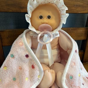 May include: A vintage baby doll with a pacifier, wearing a bonnet and wrapped in a pink blanket with colorful polka dots. The doll has large brown eyes and is sitting on a wooden chair.