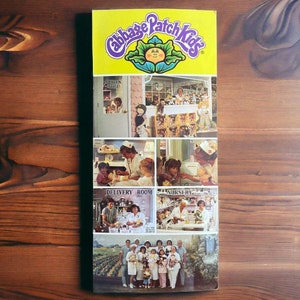 May include: A colorful brochure for Cabbage Patch Kids dolls. The brochure features a yellow cover with the Cabbage Patch Kids logo and images of dolls in various settings, including a delivery room, a nursery, and a store.