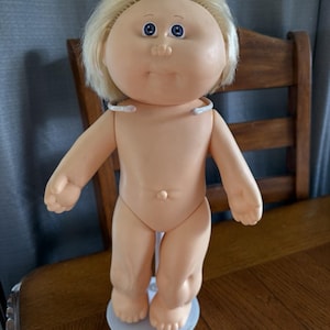 Peut inclure: Une poupée Cabbage Patch Kid vintage avec une peau de couleur pêche, des yeux bleus et des cheveux blonds courts et clairs. La poupée est debout sur un support blanc et a une expression neutre. La poupée mesure environ 36 cm de haut.
