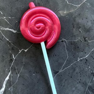 May include: A large, swirled, red lollipop with a light blue stick. The candy has a spiral design and a glossy finish. The lollipop is set against a dark, marbled background.