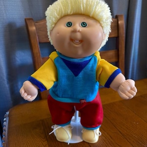May include: A vintage Cabbage Patch Kid doll with blonde yarn hair, green eyes, and a smiling expression. The doll is wearing a blue and yellow top, red pants, and yellow shoes. The doll is standing on a white stand.