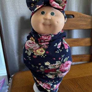 May include: A Cabbage Patch Kids doll with a round face, green eyes, and a dimple. The doll is wrapped in a navy blue floral fabric and wears a matching bow. The doll is on a white plate.