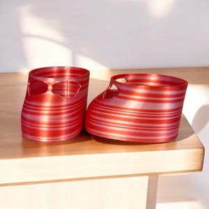 May include: Two red and white striped decorative glass containers shaped like baby shoes. The containers have a rounded shape with a cut-out design on the top and a flat base. They are sitting on a light-colored wooden surface.