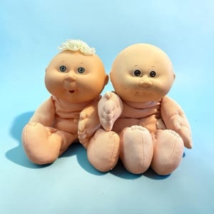 Puede incluir: Dos muñecas Cabbage Patch Kids vintage. Una muñeca tiene un mechón de pelo rubio. Ambas muñecas tienen ojos azules, mejillas rosadas y visten trajes de tela color melocotón. Las muñecas están sentadas sobre un fondo azul claro.