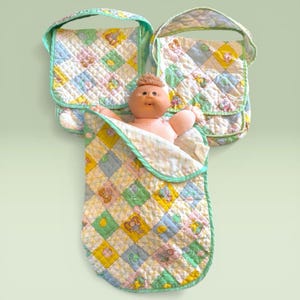 Puede incluir: Una muñeca vintage con una tez rosada suave, acurrucada en una manta acolchada. La manta y dos bolsos a juego presentan un diseño de patchwork pastel con personajes de dibujos animados. Los bolsos tienen ribetes y asas verde menta.
