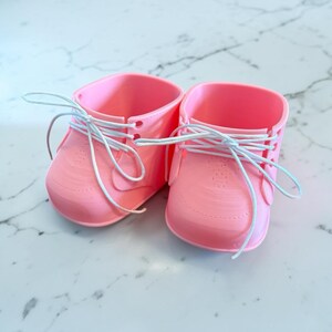 May include: A pair of pink baby shoes with white laces. The shoes have a classic sneaker design, with a rounded toe and small perforations. The laces are tied in a bow. The shoes are made of a smooth material.