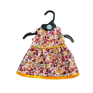 May include: A sleeveless dress with a colorful leaf pattern in shades of orange, red, and purple, trimmed with yellow. The dress is hanging on a black hanger, showcasing its design and details.