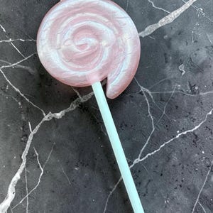Può includere: Un grande lecca-lecca rosa con un design a spirale e un bastoncino azzurro. La caramella ha una lucentezza perlescente, che le conferisce un aspetto scintillante. Il lecca-lecca è posto su uno sfondo scuro e marmorizzato.