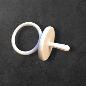 Pode incluir: Um objeto branco e minimalista em forma de chupeta. O design apresenta um anel circular conectado a um disco plano e circular com uma alça cilíndrica curta. O objeto está sobre um fundo escuro e texturizado.